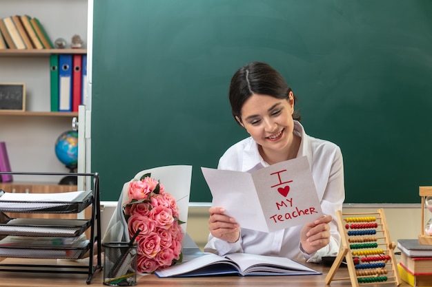 Smiling young female teacher holding and reading greeting card sitting at table with school tools in classroom