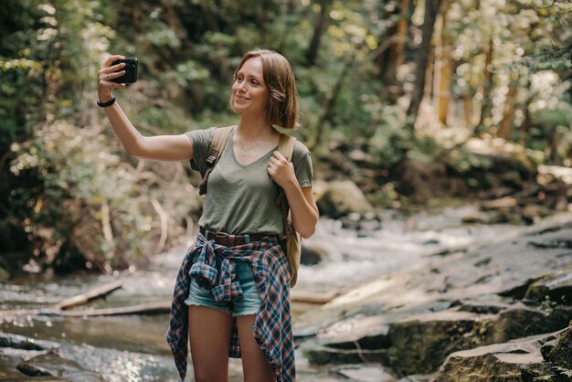 Smiling young female traveler with backpack making selfie while standing by the mountain river