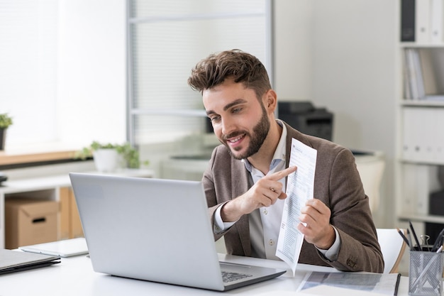 Smiling young financial consultant with beard talking to taxpayer via videoconferencing app on laptop while explaining how to fill tax papers