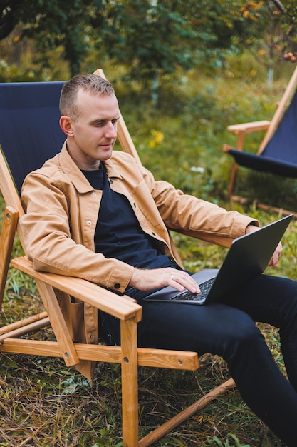 Smiling young freelancer man sitting in wooden chair with laptop while working outdoors in garden Remote work