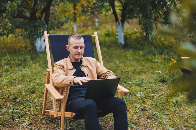 Smiling young freelancer man sitting in wooden chair with laptop while working outdoors in garden Remote work