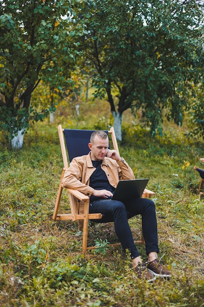 Smiling young freelancer man sitting in wooden chair with laptop while working outdoors in garden Remote work