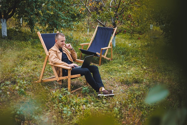 Smiling young freelancer man sitting in wooden chair with laptop while working outdoors in garden Remote work