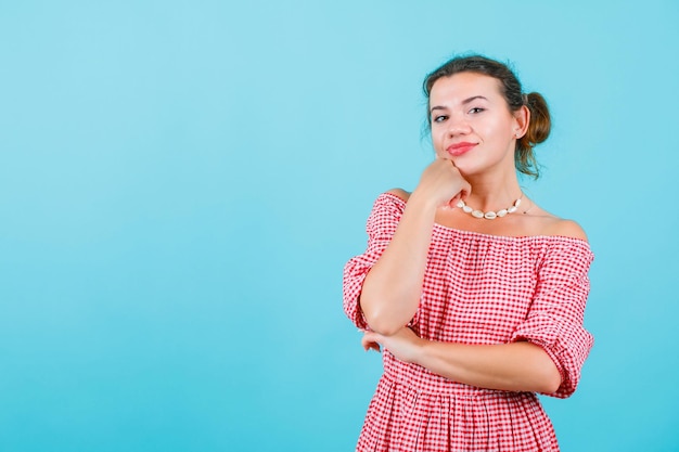 Smiling young girl is looking at camera by holding fist on chin on blue background