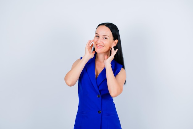 Smiling young girl is posing to camera by holding hands near ears on white background