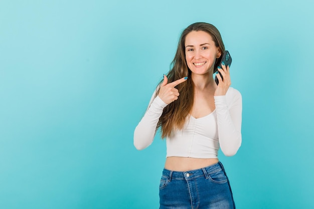 Smiling young girl is showing mobile in hand with forefinger on blue background