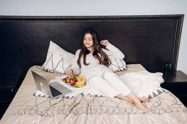 Smiling young girl lies in bed after work looks funny movie on her laptop and eats fruit