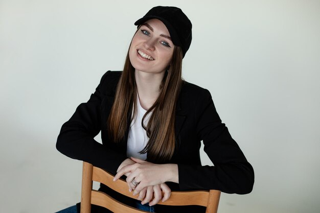 Photo smiling young girl sitting on chair gray studio background copy space female casual black blazer and cap trendy clothes and headdresses monochromatic collection of fashionable clothing for women