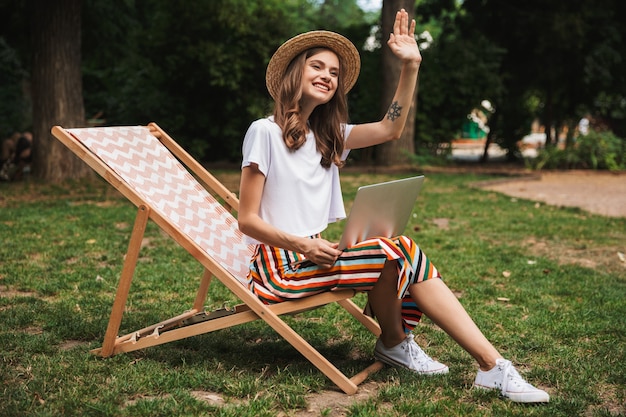 Smiling young girl sitting with laptop computer at the park outdoors, waving