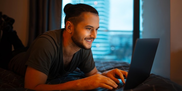 Smiling young guy lying on bed and working home on laptop Typing on keyboard