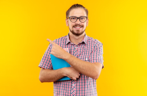 Smiling young guy student wearing backpack with glasses holding book points at side isolated on orange wall with copy space