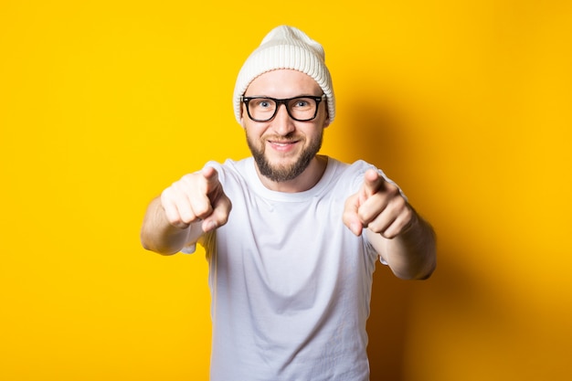 Smiling young guy with a beard in a cap and glasses with index fingers points forward.