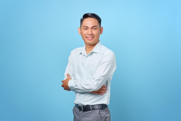 Smiling young handsome businessman crossing hands and look at camera on blue background