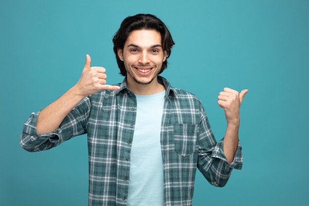 smiling young handsome man looking at camera showing call gesture pointing to side isolated on blue background