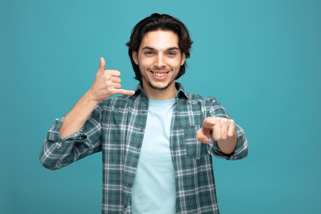 smiling young handsome man looking and pointing at camera showing call gesture isolated on blue background