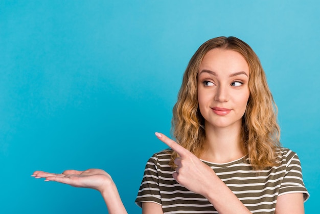 Smiling young lady holding hand with open palm advertising showing free empty space poiting isolated on blue studio wall