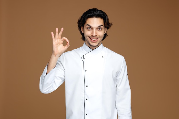 smiling young male chef wearing uniform looking at camera showing ok sign isolated on brown background