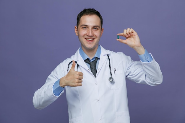 Smiling young male doctor wearing medical robe and stethoscope around neck showing capsule looking at camera showing thumb up isolated on purple background