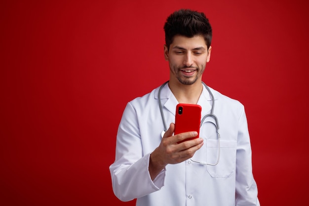 Smiling young male doctor wearing medical uniform and stethoscope around his neck holding and looking at mobile phone isolated on red background with copy space