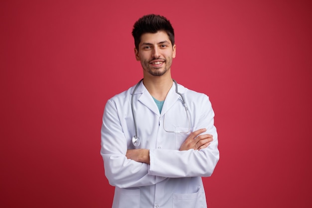 Smiling young male doctor wearing medical uniform and stethoscope around his neck keeping arms crossed looking at camera isolated on red background