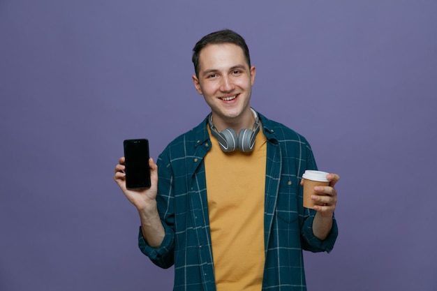 Smiling young male student wearing headphones around neck holding paper coffee cup looking at camera showing mobile phone isolated on purple background