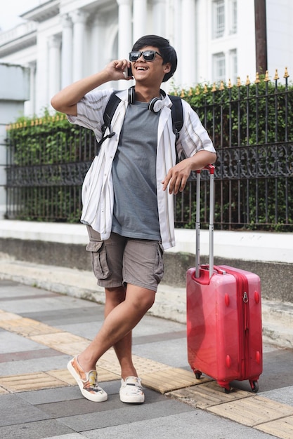 Smiling young male traveler in casual style bring suitcase make a call at pedestrian walk