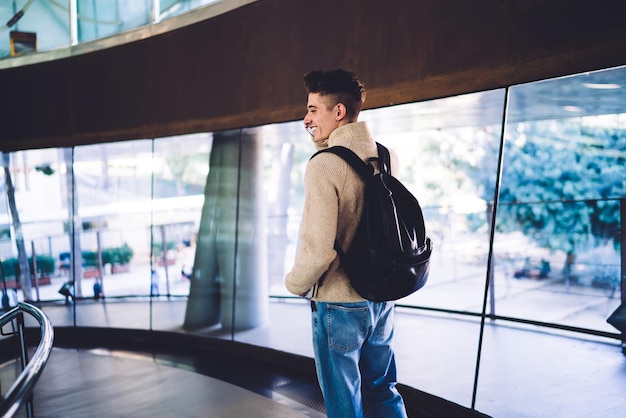 Smiling young male with backpack talking on phone near window