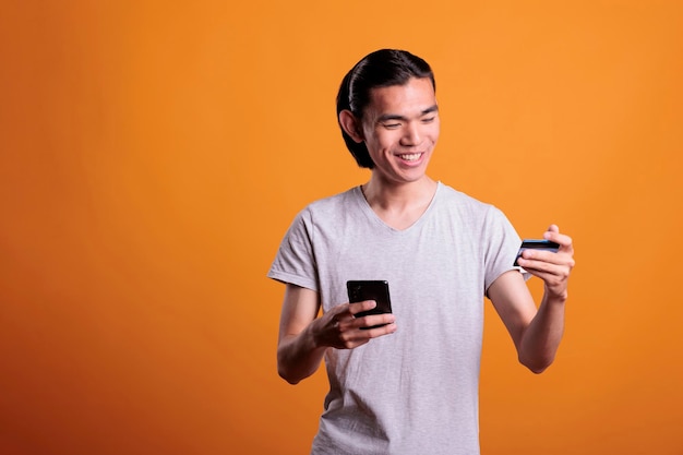 Smiling young man buying products in internet shop, making order using smartphone app. Asian teenager holding debit card and searching goods in online store, e commerce concept