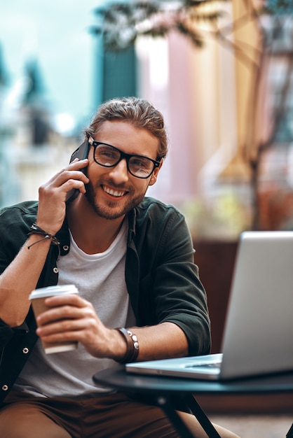 Smiling young man in casual clothing talking on the smart phone and using laptop while working in cafe