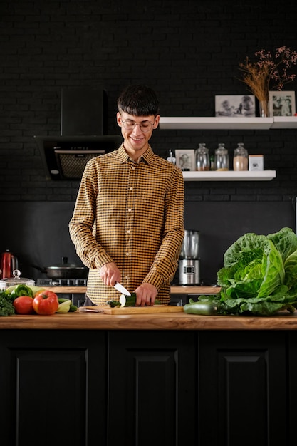 Smiling young  man cooking salad, cutting fresh vegetables in modern black kitchen