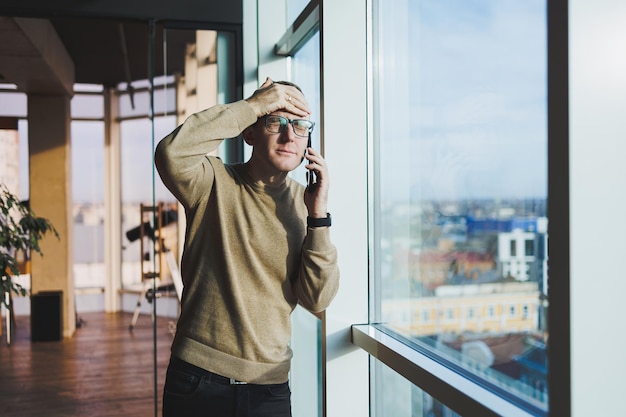 A smiling young man in glasses is smiling and talking on the phone on a mobile phone while relaxing in the office A young freelancer works remotely