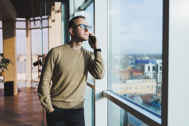 A smiling young man in glasses is smiling and talking on the phone on a mobile phone while relaxing in the office A young freelancer works remotely