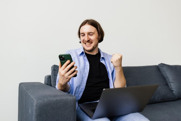 Smiling young man holding mobile phone while sitting on a sofa at home with laptop and celebrating