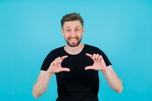 Smiling young man is showing size gesture with hands on blue background