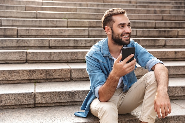 Smiling young man sitting on stairs outdoors, using mobile phone