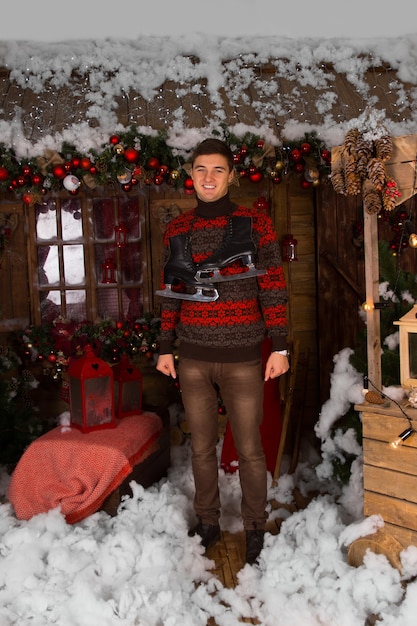 Smiling Young Man Standing at the House with Plenty Christmas Decors in Winter Fashion Outfit, with Ice Skates Hanging Around his Neck, While Looking at the Camera.