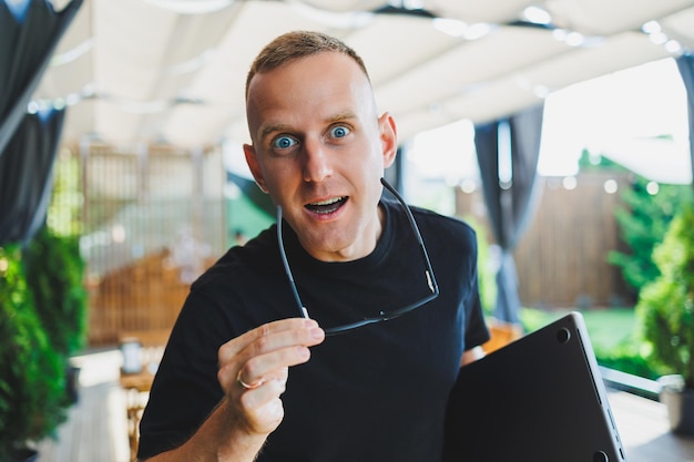 A smiling young man stands on a summer terrace in a cafe with a laptop in his hands Work online freelancer Remote summer job online