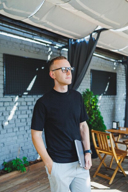 A smiling young man stands on a summer terrace in a cafe with a laptop in his hands Work online freelancer Remote summer job online