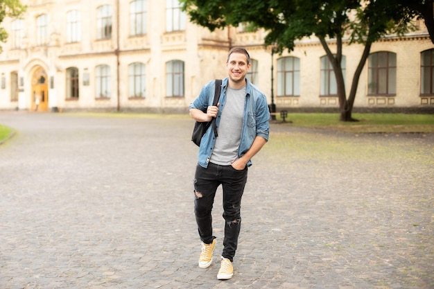 Smiling young man with backpack stand against university on the background in park