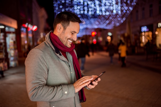 Smiling young man with mobile phone on the street with Christmas decoration.