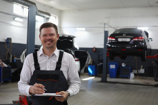 Smiling young mechanic writes on clipboard machines in background