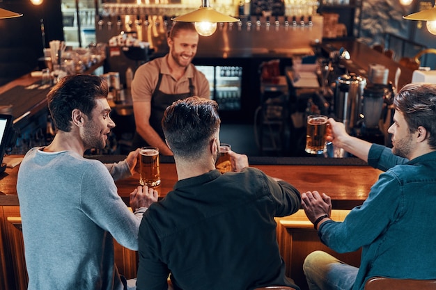 Smiling young men in casual clothing drinking beer while sitting in the pub together