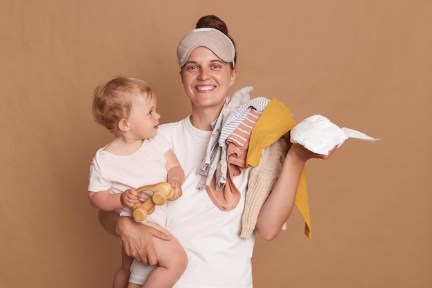 Smiling young mother with bun hairstyle holding her toddler daughter clothing and diaper looking at camera with toothy smile enjoying her maternity leave posing isolated over brown background