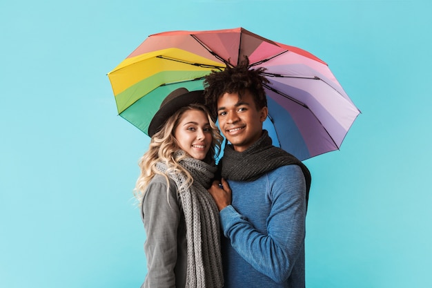 Smiling young multiracial couple wearing scarves standing with an umbrella isolated over blue wall