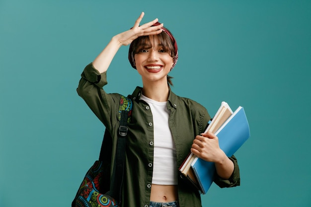 Smiling young student girl wearing bandana and backpack holding note pads with pen keeping hand on forehead looking at camera into distance isolated on blue background