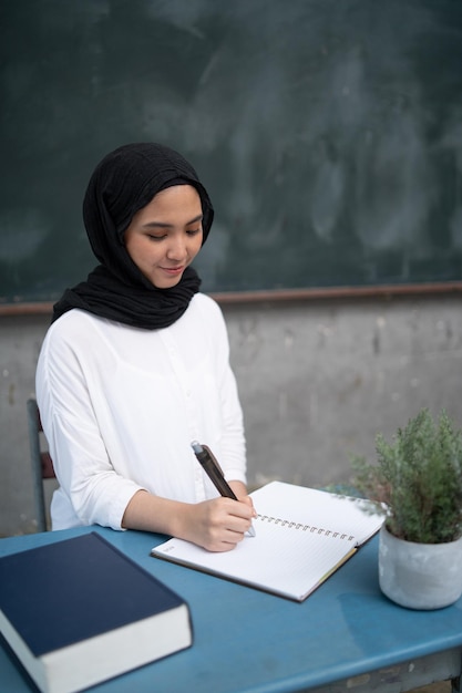 Photo smiling young teacher writing in book while sitting on table at classroom