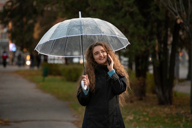 Smiling Young woman in autumn park with transparent umbrella talking on phone Girl walking in the park