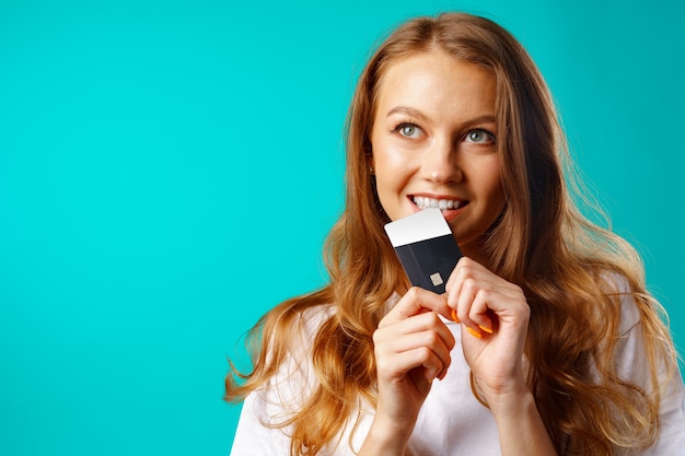 Smiling young woman biting a credit card within temptation of shopping against blue background