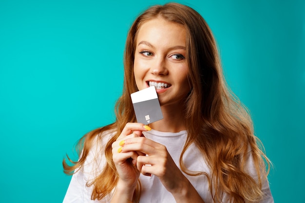 Smiling young woman biting a credit card within temptation of shopping against blue background