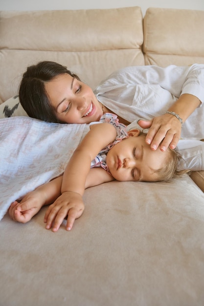 Smiling young woman caressing adorable baby sleeping on comfortable sofa in living room at home